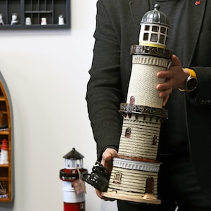 May include: A white and brown ceramic lighthouse model with a black roof and windows. The lighthouse is on multiple tiers and has a yellow band around the top. The model is being held by a person wearing a black jacket.