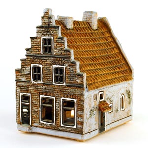 Può includere: Una casa in miniatura in ceramica dettagliata con facciata in mattoni, tetto in tegole e più finestre. La casa ha un esterno di colore chiaro con un bagliore caldo e invitante che emana dall'interno. Il design generale ricorda un edificio europeo tradizionale.