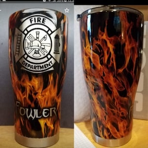 Op de afbeelding: Een zwarte en oranje roestvrijstalen beker met een vuurontwerp. De beker heeft een zilveren deksel en een zilveren basis. De voorkant van de beker heeft een zilveren embleem met de woorden "Fire Department" en "Fowler" erop gedrukt.