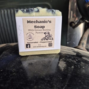May include: A rectangular bar of soap with a marbled black and off-white top. A beige paper label features the text Mechanic's Soap With Grease Busting Pumice, Spearmint + Peppermint, 5 oz., and a graphic of a person holding a wrench.