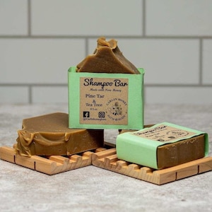 May include: Three bars of shampoo soap on wooden soap dishes. The soap is brown and the wrappers are green with white text that reads "Shampoo Bar Made with Pure Honey Pine Tar & Tea Tree 4.5 oz. Carlin Honey Bath & Body @Carlinhoneybees".