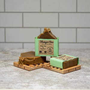 May include: Three brown shampoo bars with light green paper labels. The labels feature the text Shampoo Bar, Made with Pure Honey, Pine Tar & Tea Tree, 4.2 oz, and Carlin Honey Bath & Body. The bars are resting on two light-colored wooden soap dishes.
