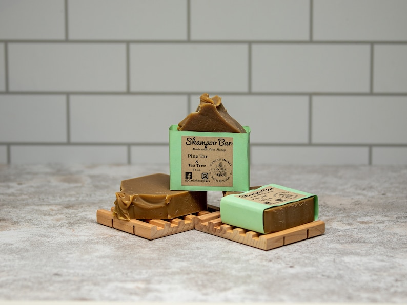 May include: Three brown shampoo bars with light green paper labels. The labels feature the text Shampoo Bar, Made with Pure Honey, Pine Tar & Tea Tree, 4.2 oz, and Carlin Honey Bath & Body. The bars are resting on two light-colored wooden soap dishes.