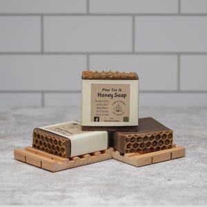 May include: Three bars of Pine Tar & Honey Soap, each with a honeycomb design, rest on small wooden soap dishes. The soap bars have a light brown color with a white label that reads "Pine Tar & Honey Soap". The background is a white tile wall.