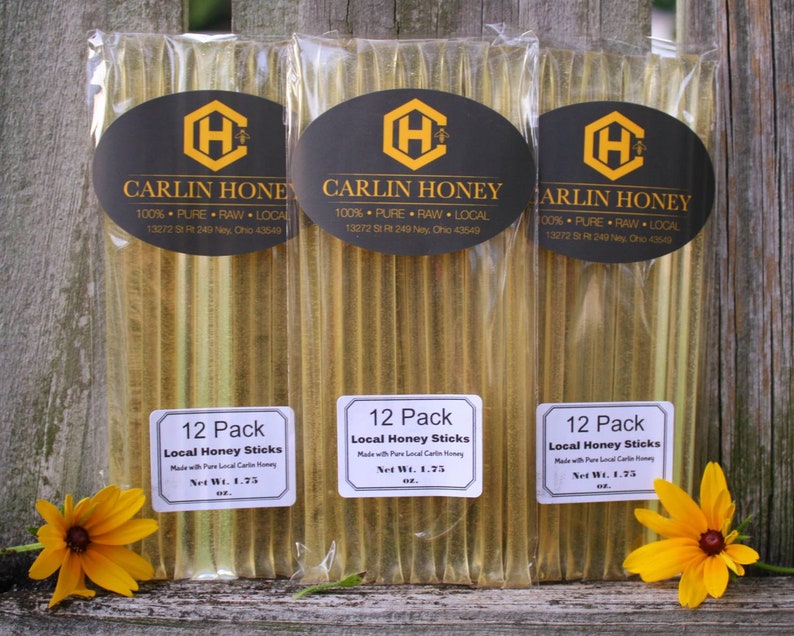 Puede incluir: Tres paquetes de 12 palitos de miel, cada uno etiquetado con "12 Pack Local Honey Sticks" y "Carlin Honey 100% Pure Raw Local 13272 St Rt 249 Ney, Ohio 43549". Los palitos de miel son transparentes y est&aacute;n llenos de miel. Los paquetes est&aacute;n sobre una superficie de madera con una flor amarilla delante de cada paquete.