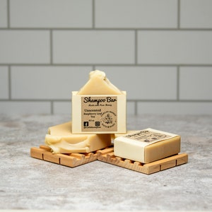 May include: Two rectangular beige shampoo bars resting on wooden soap dishes. The upright bar features a label reading Shampoo Bar, Made with Pure Honey, Unscented, Raspberry Leaf Tea, 4.5 oz, and Carlin Honey Bath & Body with social media icons.