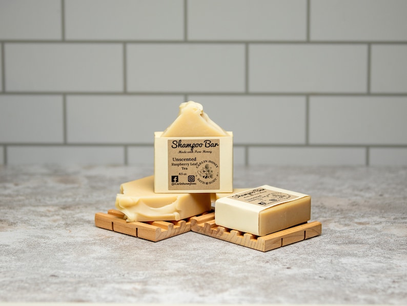 May include: Two rectangular beige shampoo bars resting on wooden soap dishes. The upright bar features a label reading Shampoo Bar, Made with Pure Honey, Unscented, Raspberry Leaf Tea, 4.5 oz, and Carlin Honey Bath & Body with social media icons.