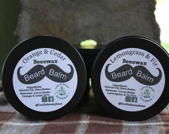 Può includere: Due barattoli rotondi neri con etichette bianche. Le etichette hanno un grafico di baffi neri e il testo "Beeswax Beard Balm". Il barattolo a sinistra ha il testo "Orange & Cedar" e il barattolo a destra ha il testo "Lemongrass & Fir". Entrambi i barattoli hanno un elenco di ingredienti e il testo "@Carlinhoneybees".