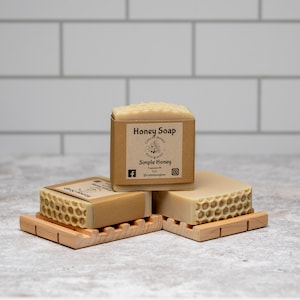 May include: Three bars of honey soap on wooden soap dishes. The top bar has a label that reads "Honey Soap" and "Simple Honey".