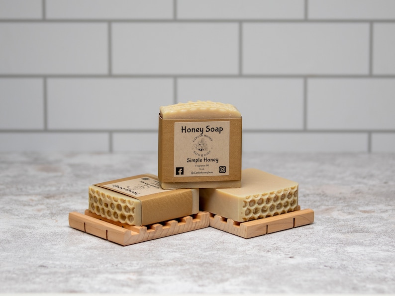 May include: Three bars of honey soap on wooden soap dishes. The top bar has a label that reads "Honey Soap" and "Simple Honey".
