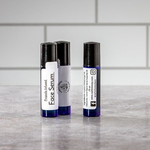 May include: Three blue glass roller bottles with white labels. The labels read "Propolis Infused Face Serum" and include the brand name "Carlin Honey" and social media icons.