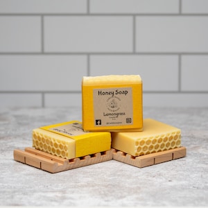 May include: Three bars of yellow honey soap with a honeycomb pattern on the top. The soap is labeled "Honey Soap" and "Lemongrass" with a small bee illustration. The soap is on a wooden soap dish.