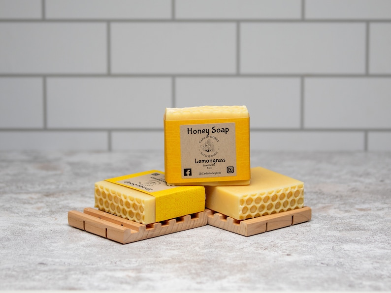 May include: Three bars of yellow honey soap with a honeycomb pattern on the top. The soap is labeled "Honey Soap" and "Lemongrass" with a small bee illustration. The soap is on a wooden soap dish.