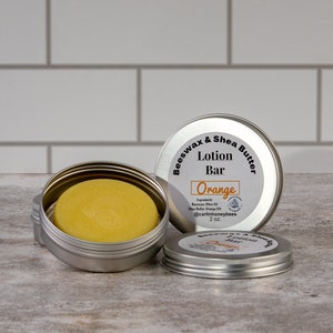 May include: A silver tin with a lid containing a round, yellow lotion bar. The tin is labelled "Beeswax & Shea Butter Lotion Bar Orange" with the ingredients listed below. The tin is 2 oz.