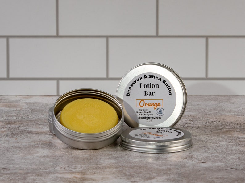 May include: A silver tin with a lid containing a round, yellow lotion bar. The tin is labelled "Beeswax & Shea Butter Lotion Bar Orange" with the ingredients listed below. The tin is 2 oz.