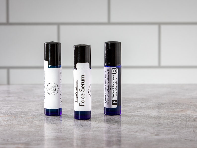 May include: Three small blue glass bottles with black roll-on tops. Each bottle has a white label with black text that reads "Propolis Infused Face Serum". The label on the right bottle also includes the text "Ingredients: Propolis Infused, Chamomile, Carrot Seed &  Merlot Essential Oil. .75 oz. www.carlinhoney.com @carlinhoneybees".