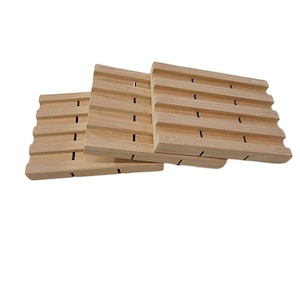 May include: Three wooden soap dishes with raised ridges and drainage holes. The dishes are made of light brown wood and are stacked on top of each other.