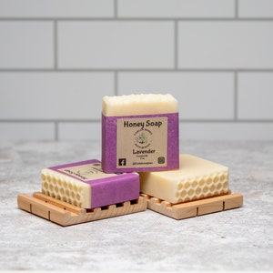May include: Three bars of honey soap with a lavender scent. The top bar has a label that reads "Honey Soap" and "Lavender". The bars are on wooden soap dishes.