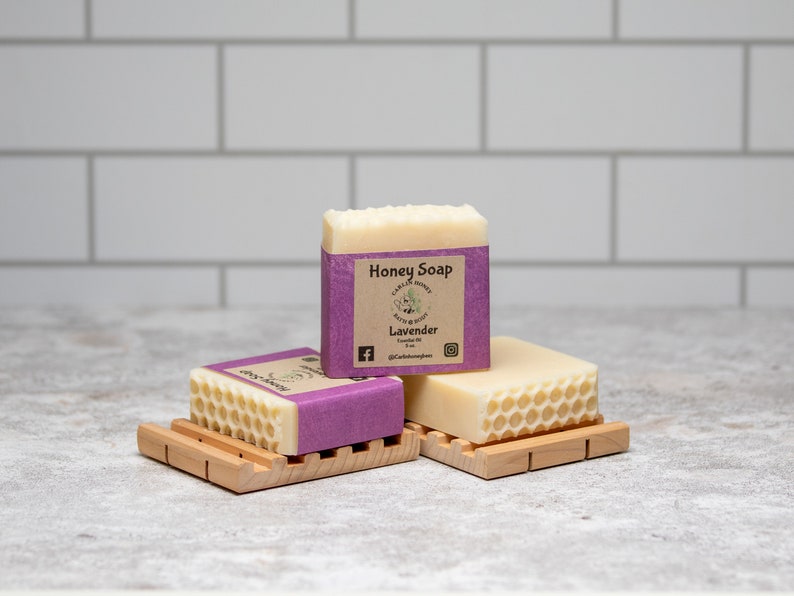 May include: Three bars of honey soap with a lavender scent. The top bar has a label that reads "Honey Soap" and "Lavender". The bars are on wooden soap dishes.