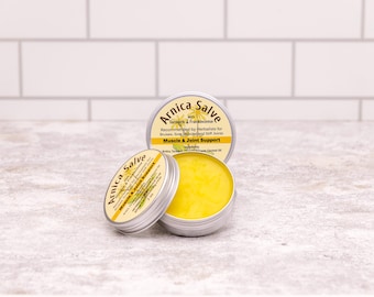 Puede incluir: Tres pequeños botes plateados de Arnica Salve con etiquetas amarillas y un bálsamo amarillo brillante en el interior de uno. Las etiquetas dicen "Arnica Salve" con texto sobre ingredientes y usos, incluyendo "Muscle & Joint Support".