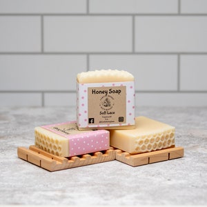 May include: Three bars of honey soap with a pink polka dot design and a label that reads "Honey Soap" and "Soft Lace". The soap bars are on a wooden soap dish.