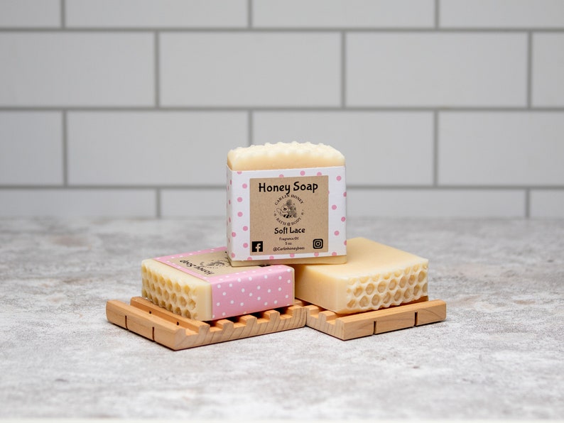 May include: Three bars of honey soap with a pink polka dot design and a label that reads "Honey Soap" and "Soft Lace". The soap bars are on a wooden soap dish.