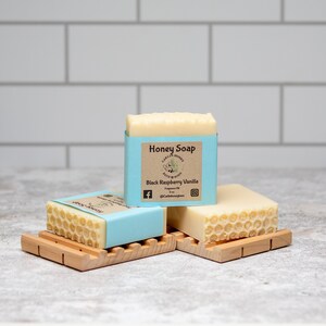 May include: Three bars of honey soap on wooden soap dishes. The soap is wrapped in blue paper with a label that reads "Honey Soap" and "Black Raspberry Vanilla".