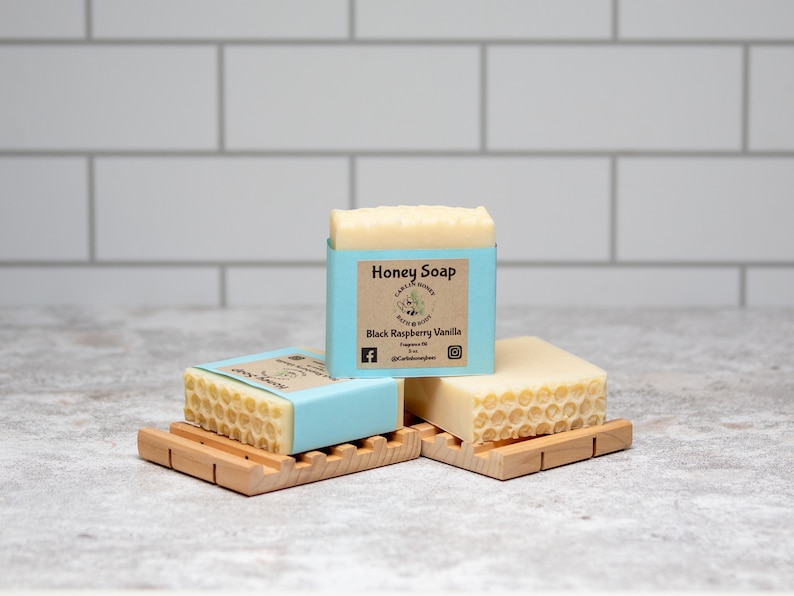 May include: Three bars of honey soap on wooden soap dishes. The soap is wrapped in blue paper with a label that reads "Honey Soap" and "Black Raspberry Vanilla".