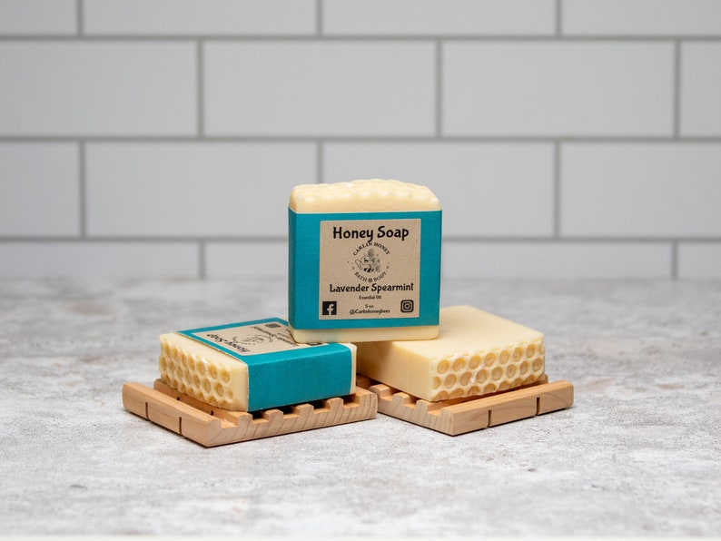May include: Three bars of honey soap with a blue and white label that reads "Honey Soap" and "Lavender Spearmint". The soap bars are on wooden soap dishes.