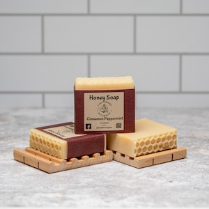 May include: Three bars of honey soap with a honeycomb design. The top bar has a brown label that reads "Honey Soap" and "Cinnamon Peppermint".