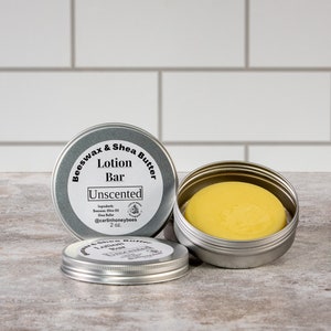 May include: A silver tin with a label that reads "Beeswax & Shea Butter Lotion Bar Unscented" and contains a yellow lotion bar. The tin is 2 oz.