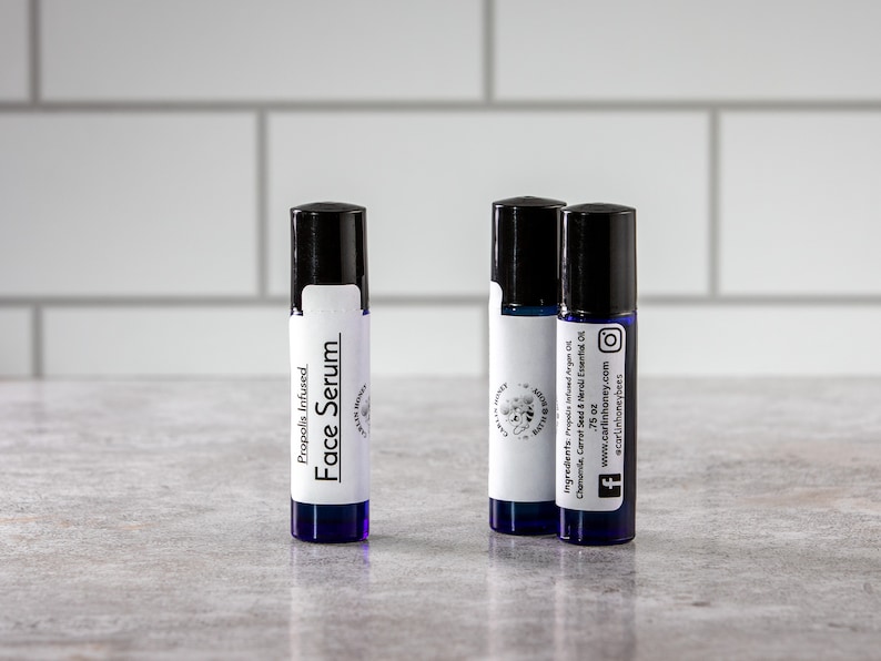 May include: Three small blue glass bottles with black roll-on tops and white labels. The labels have black text that reads "Propolis Infused Face Serum" and "Ingredients: Propolis Infused Argan Oil, Chamomile, Carrot Seed & Neroli Essential Oil. 0.75 oz. www.carlinhoney.com #acarlinhoneybees".
