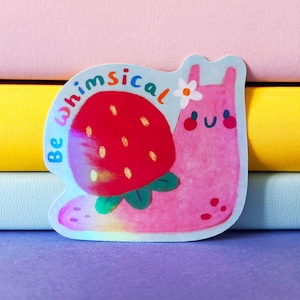 May include: A whimsical sticker depicting a pink snail with a strawberry shell and a small white flower. The sticker has the words "Be whimsical" in blue. The background is a stack of colourful books.