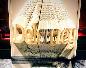 Puede incluir: Una escultura de arte de libro con el nombre "Delaney" plegado en las páginas. El libro está de pie sobre una superficie blanca con una cinta negra atada alrededor de la parte inferior.