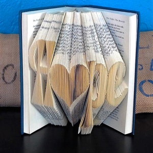 May include: A book art sculpture of the word "Hope" created by folding the pages of a book. The book is open and the pages are folded to create the letters. The book is on a dark surface.