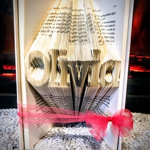 May include: A book folded into the shape of the word "Olivia" with a pink ribbon tied around it. The book is sitting on a white fuzzy surface.