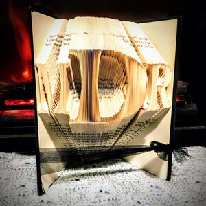 May include: A book art sculpture of the word "LOVE" created by folding the pages of a book. The sculpture is displayed on a white surface with a black ribbon.