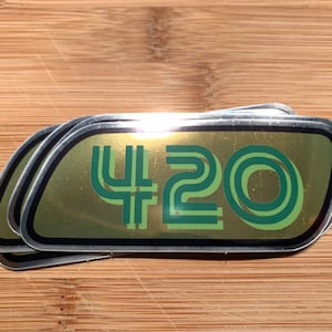 May include: A stack of three gold-coloured stickers with a green 420 design. The stickers have a silver border and a slightly rounded rectangular shape.