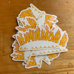 Może przedstawiać: Stos białych naklejek z ż&oacute;łto-czerwonym wzorem płomieni. Tekst "BAVAMOW" jest napisany ż&oacute;łtą, pogrubioną czcionką z czerwonym obramowaniem. Tekst "de coraz&oacute;n" jest napisany mniejszą, czarną czcionką pod gł&oacute;wnym tekstem.