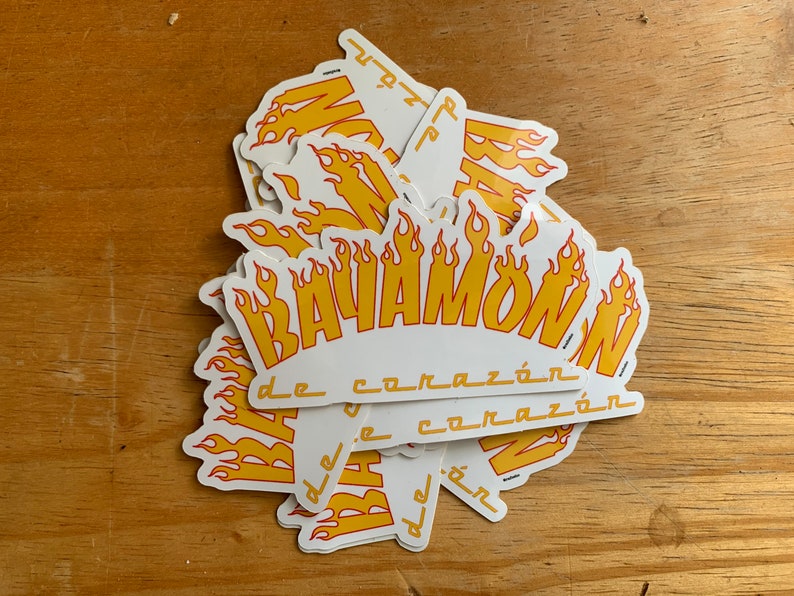 Może przedstawiać: Stos białych naklejek z ż&oacute;łto-czerwonym wzorem płomieni. Tekst "BAVAMOW" jest napisany ż&oacute;łtą, pogrubioną czcionką z czerwonym obramowaniem. Tekst "de coraz&oacute;n" jest napisany mniejszą, czarną czcionką pod gł&oacute;wnym tekstem.