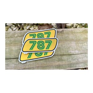 Peut inclure: Trois autocollants jaunes et verts avec le num&eacute;ro 787 imprim&eacute; dessus. Les autocollants sont empil&eacute;s les uns sur les autres.