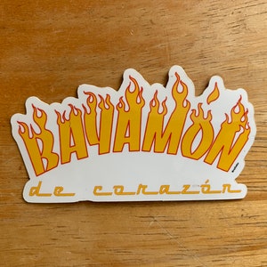 Może przedstawiać: Biała naklejka z napisem "BAYAMON" w ż&oacute;łtych literach z czerwonymi obramowaniami. Litery są otoczone ż&oacute;łtymi płomieniami. Tekst "de coraz&oacute;n" jest napisany mniejszymi literami pod słowem "BAYAMON".