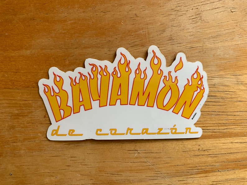 Może przedstawiać: Biała naklejka z napisem "BAYAMON" w ż&oacute;łtych literach z czerwonymi obramowaniami. Litery są otoczone ż&oacute;łtymi płomieniami. Tekst "de coraz&oacute;n" jest napisany mniejszymi literami pod słowem "BAYAMON".