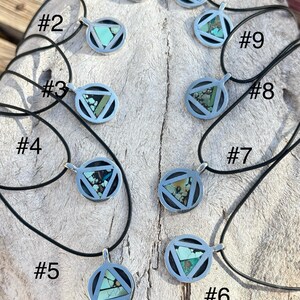 Puede incluir: Una colecci&oacute;n de diez collares con colgantes de plata con cordones negros. Cada colgante presenta un marco circular con una incrustaci&oacute;n triangular de turquesa y otras variaciones de piedra. Los collares se exhiben sobre una superficie clara y texturizada.