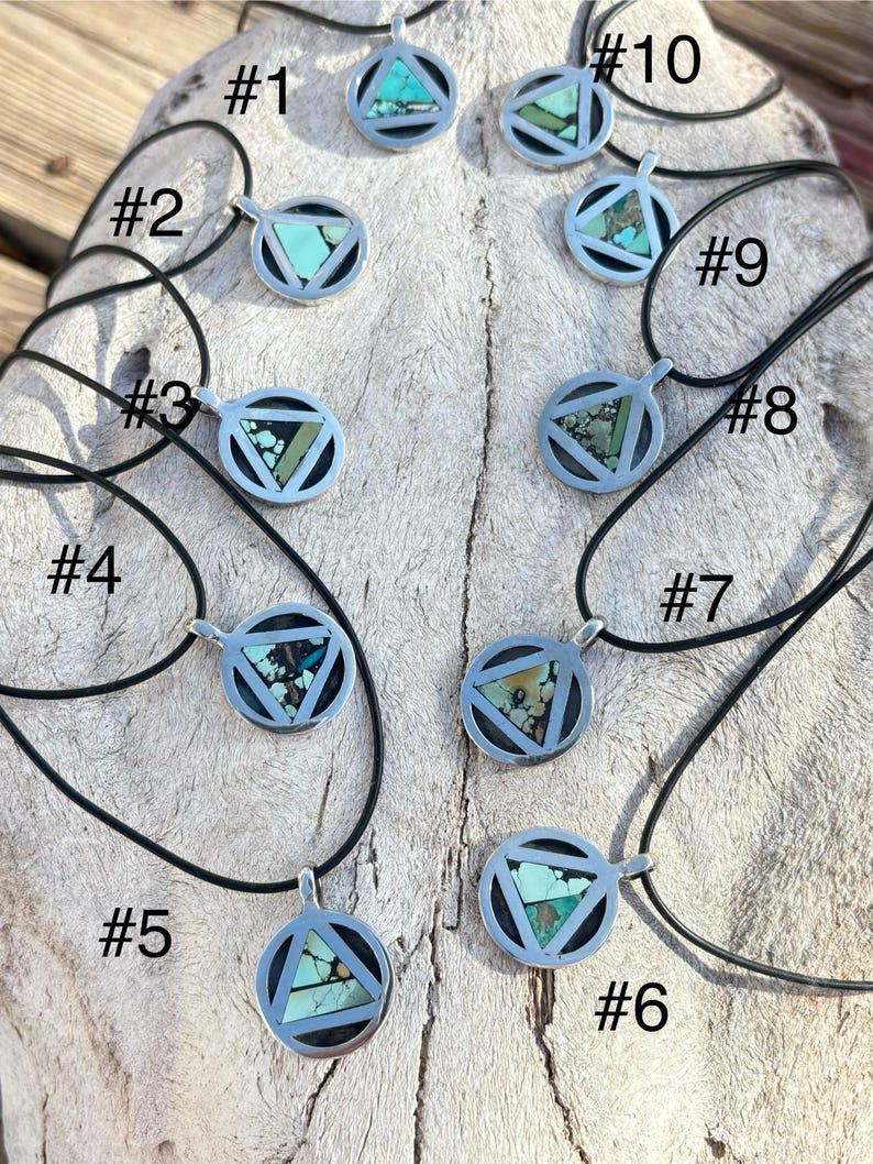 Puede incluir: Una colecci&oacute;n de diez collares con colgantes de plata con cordones negros. Cada colgante presenta un marco circular con una incrustaci&oacute;n triangular de turquesa y otras variaciones de piedra. Los collares se exhiben sobre una superficie clara y texturizada.