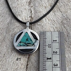 Puede incluir: Colgante de plata con incrustaciones de piedra turquesa, con un dise&ntilde;o de tri&aacute;ngulo dentro de un c&iacute;rculo, colgado de un cord&oacute;n negro. El colgante mide aproximadamente 2,5 cm de di&aacute;metro. El texto "#1" es visible debajo.