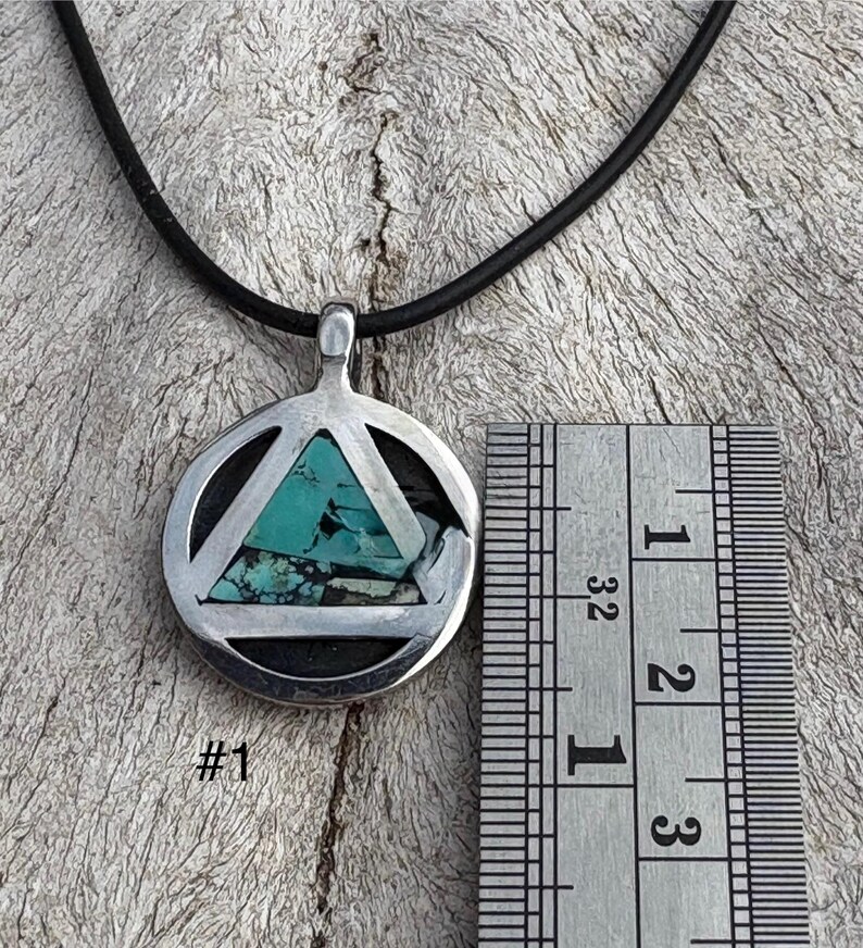 Puede incluir: Colgante de plata con incrustaciones de piedra turquesa, con un dise&ntilde;o de tri&aacute;ngulo dentro de un c&iacute;rculo, colgado de un cord&oacute;n negro. El colgante mide aproximadamente 2,5 cm de di&aacute;metro. El texto "#1" es visible debajo.