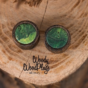 May include: Two round wooden ear plugs with a dark brown border and a green, marbled center. The plugs are set on a wooden surface. The text "Woody WoodPlugs - est. 2015" is visible below the plugs.