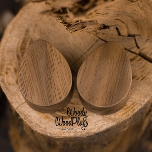May include: Two brown wooden ear gauges, shaped like teardrops, are displayed on a wooden surface. The gauges are made by "Woody Wood Plugs" est. 2015.