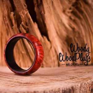 May include: A red and black wooden ring with a smooth, polished finish. The ring is sitting on a wooden surface. The text "Woody WoodPlugs - est. 2015" is visible in the background.